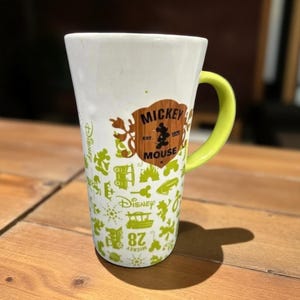 May include: A white ceramic Mickey Mouse mug with a lime green handle. The mug features a brown emblem with "Mickey Mouse" and the year "1928". The mug is decorated with green Disney-themed graphics and the number "82".