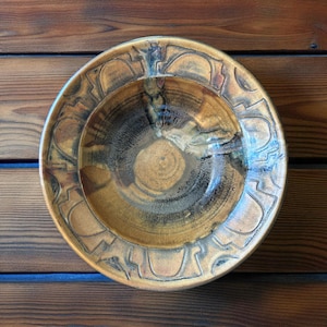 Peut inclure: Un bol en céramique fait à la main avec un large bord et un centre profond. Le bol présente une palette de couleurs marron, beige et noire avec une finition brillante. Le bord est orné d'un motif géométrique répétitif. Le bol est probablement utilisé pour servir des aliments.