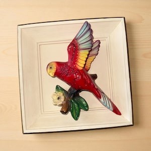 May include: A vibrant red parrot sculpture with yellow, blue, and green accents, perched on a branch with a white flower and green leaves. The bird is set against a square, off-white background with a dark brown border, creating a decorative wall art piece.
