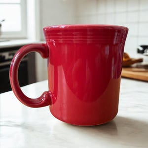 Peut inclure: Une tasse en céramique rouge vif avec une anse incurvée. La tasse a une forme cylindrique avec un bord légèrement évasé et une finition brillante. La tasse est posée sur une surface en marbre blanc.