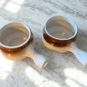 Puede incluir: Dos cuencos de sopa de cerámica con asas. Los cuencos tienen un esquema de color degradado, que va desde un beige claro en la base hasta un marrón intenso en el borde. Las asas son de color beige claro. Los cuencos están vacíos y listos para ser llenados.