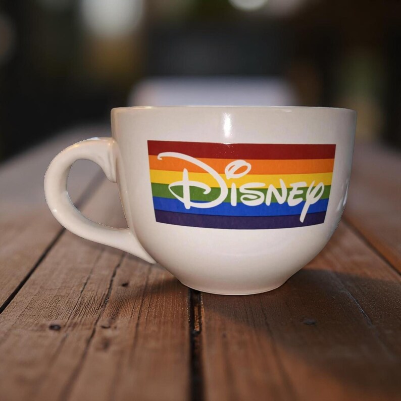 May include: White ceramic mug with a large handle. The mug features the Disney logo in a rainbow design. The mug is sitting on a wooden surface.