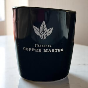 May include: A black ceramic coffee mug with the Starbucks logo and the words "COFFEE MASTER" in white. The mug has a glossy finish and a slightly angled design. The logo features a stylized leaf and coffee bean design.
