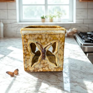 May include: A square, textured, ceramic container with a butterfly cutout design. The container is mainly tan with brown accents and a light-coloured rim. A butterfly rests on the surface nearby. The container is likely for decorative purposes.