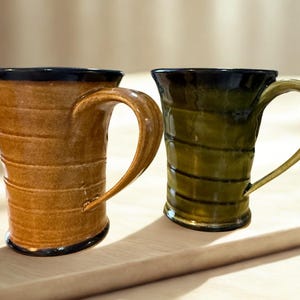 May include: Two handcrafted ceramic mugs. One is a warm brown with a dark blue rim and handle, the other is olive green with a black rim and handle. Both mugs have a unique, textured design and are set on a light-coloured surface.