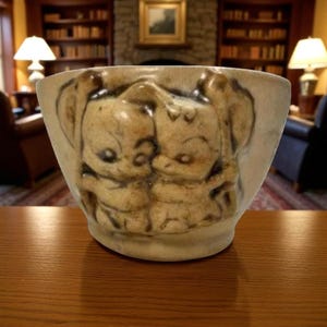 May include: A beige ceramic bowl featuring a cartoon design of two embracing figures. The figures, in light brown, have large ears. The bowl sits on a wooden surface.