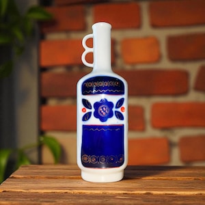 Puede incluir: Una botella de cerámica blanca con un diseño floral azul y dos asas de bucle. La botella tiene un cuello largo y un cuerpo rectangular con un patrón floral azul y detalles en rojo. La botella está sobre una superficie de madera.