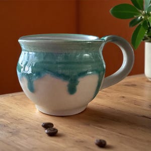 May include: A handmade ceramic mug with a rounded body and a handle. The mug features a two-tone design, with a white base and a teal-green glaze that drips down the sides. Coffee beans are scattered on the wooden surface.
