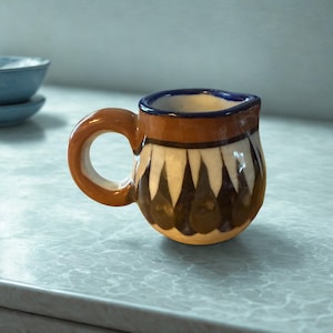 Puede incluir: Una pequeña jarra de cerámica con asa y borde marrón. El cuerpo presenta un patrón de formas blancas y marrón oscuro alternadas. El interior es azul y la jarra está sobre una superficie clara. Un cuenco azul está en el fondo.