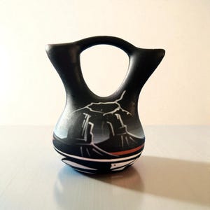 May include: Black ceramic vase with a distinctive handle and a painted landscape design. The vase has a black base with white and red stripes, and a detailed mountain scene in shades of grey and white.