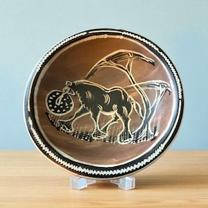 May include: A brown ceramic bowl with a black and white decorative border. The bowl features a black illustration of an animal carrying a circular object, with trees in the background. The bowl is displayed on a clear stand.