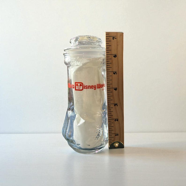May include: Clear glass jar with a lid, featuring the "Walt Disney World" logo in red. A wooden ruler is placed next to the jar, measuring approximately 7 inches in height. The jar has a unique, curved shape.