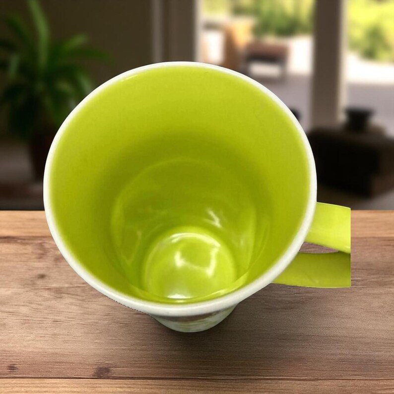May include: A lime green ceramic mug with a white rim and handle. The interior of the mug is a bright, solid green. The mug sits on a wooden surface.