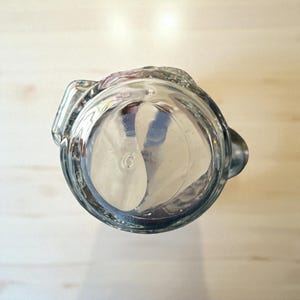 May include: Clear glass pitcher with a handle and a spout. The pitcher is empty and appears to be made of thick glass. The image is taken from a top-down perspective, showing the inside of the pitcher.