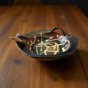 May include: A ceramic ashtray with a handle and a built-in rest. The ashtray has a black rim, a brown interior with white and black abstract designs, and a tan rest. The ashtray is on a wooden surface.