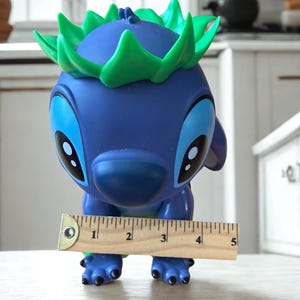Puede incluir: Una figura azul con ojos grandes y detalles verdes, que se asemeja a un personaje de dibujos animados. Una regla de madera muestra que la figura mide aproximadamente 10 cm de alto. Dise&ntilde;o juguet&oacute;n y caprichoso.