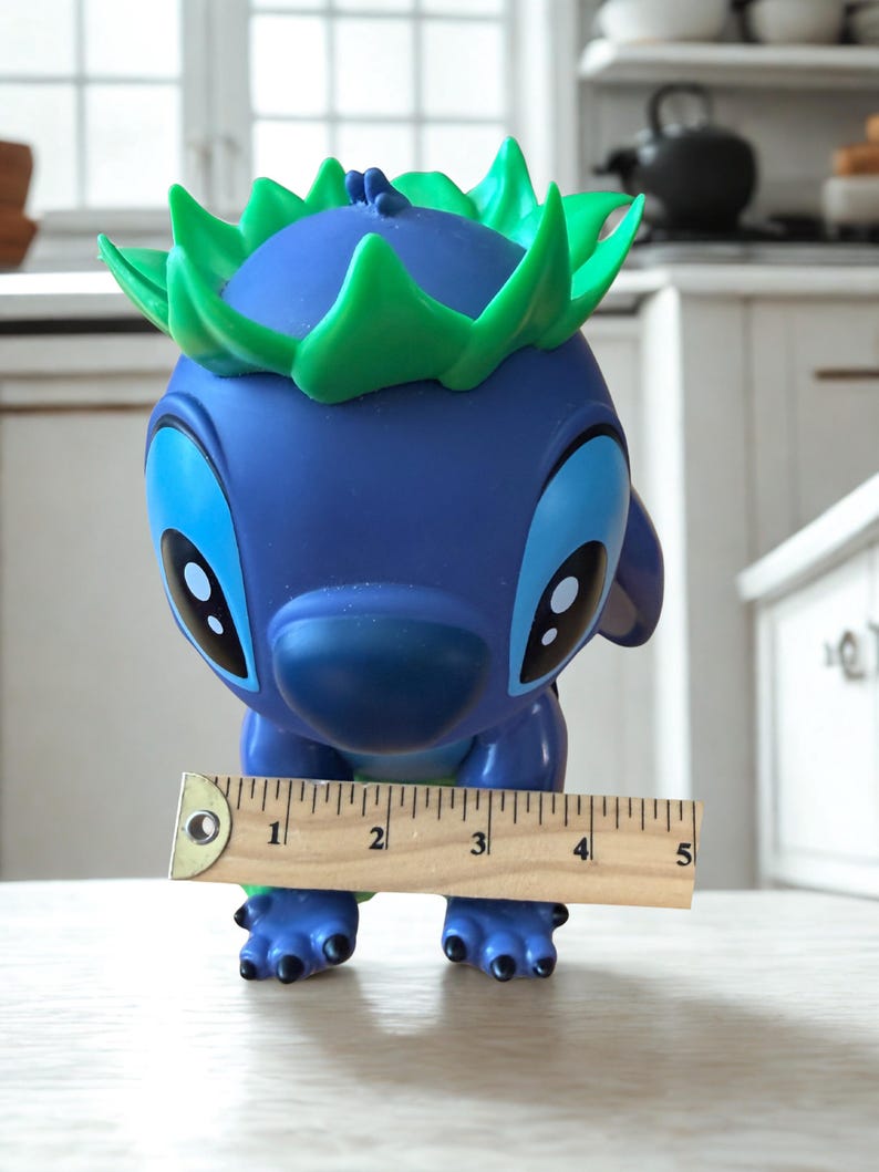 Puede incluir: Una figura azul con ojos grandes y detalles verdes, que se asemeja a un personaje de dibujos animados. Una regla de madera muestra que la figura mide aproximadamente 10 cm de alto. Dise&ntilde;o juguet&oacute;n y caprichoso.