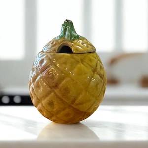 May include: A yellow ceramic pineapple-shaped container with a lid. The container has a textured surface and a green stem on top. The lid has a small opening. The container is on a white surface.