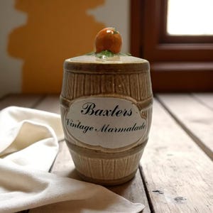 Puede incluir: Un tarro de mermelada de cerámica vintage con forma de barril, con el texto "Baxters Vintage Marmalade" en una etiqueta. La tapa está adornada con una pequeña fruta naranja. El tarro es marrón y beige, con un diseño rústico.
