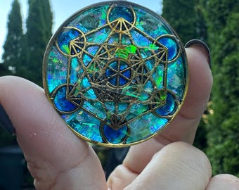 May include: A round pendant with a gold-colored frame and a geometric design. The pendant features a blue, green, and iridescent center with a Metatron's Cube design. The pendant is held by a hand against a backdrop of green foliage.
