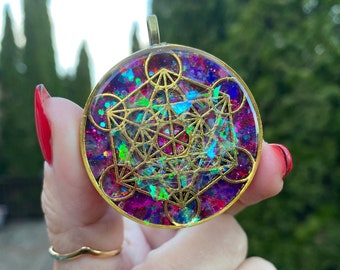 May include: A round, gold-toned pendant with a geometric design in iridescent colors, including pink, purple, blue, and green. The design is set in clear resin and has a sparkly, glittery finish.