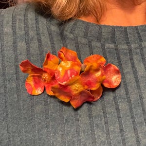 May include: A floral brooch with red, orange, and yellow petals. The brooch is made of a textured material and is pinned to a teal-colored sweater. The brooch is a decorative accessory.