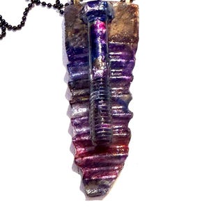 May include: A unique pendant necklace featuring a bolt design. The bolt is attached to a rectangular, textured pendant with shades of purple, blue, and red. The necklace has a black beaded chain and gold-colored clasps.