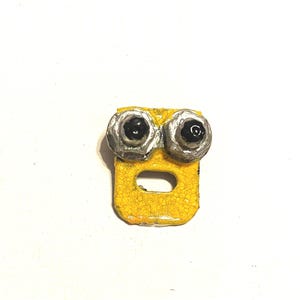 May include: A whimsical, handmade yellow object with a surprised expression. It features two large, silver-rimmed eyes with black pupils and a rectangular body with a curved opening. The object has a textured, cracked surface.