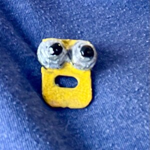 May include: A small, handmade yellow and gray charm with a cartoon-like face. The charm features two gray, round eyes with black pupils above a yellow, rectangular body with a central oval opening. The charm is set against a blue background.
