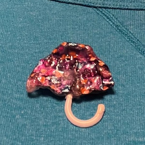 May include: A pin shaped like an umbrella with a canopy featuring a dark, multi-colored glittery finish in shades of purple, pink, and orange. The umbrella handle is a smooth, light-colored, curved hook attached to the base of the canopy.