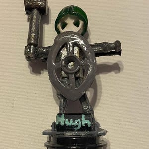 Puede incluir: Una pequeña figura de guerrero con casco verde, sosteniendo un martillo gris en una mano y un escudo gris circular en la otra. La figura se apoya sobre una base negra con el nombre Hugh pintado en letras azul claro sobre un bloque negro.