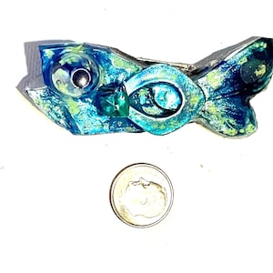 May include: A handcrafted, abstract fish-shaped brooch. The brooch features a combination of blue, green, and silver tones. The fish has a large eye and a tail fin. A dime is next to the brooch for size comparison.