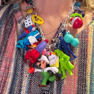 May include: A silver chain necklace adorned with a variety of colorful charms. Charms include toy figures, dice, a yellow steering wheel, a red and white bobber, and a Carl's Jr. french fries box.