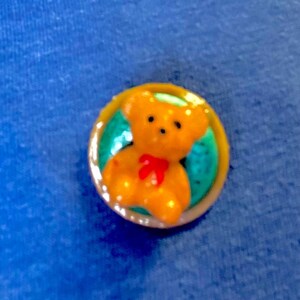 May include: A small, round button featuring a golden-yellow teddy bear with a red bow tie. The bear is set against a teal background within a gold-colored border. The button is set against a blue background.
