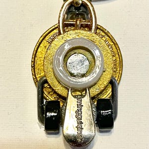 May include: A handmade pendant necklace featuring a gold-toned coin with etched details, a silver-toned charm with the text "mingguiginsu", and black accents. The pendant hangs from a black cord.