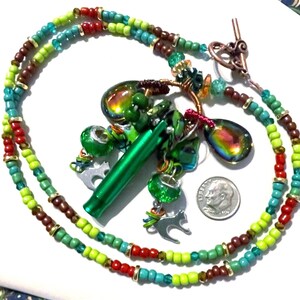 May include: A handmade necklace with a variety of beads in shades of green, brown, red, and blue. The necklace features a green pendant, iridescent glass charms, and small silver cat charms. A dime is included for size reference.
