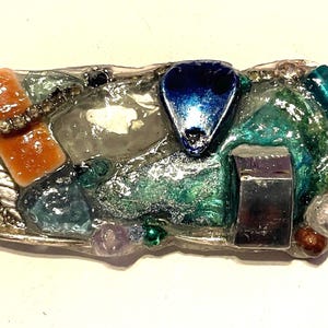 May include: A rectangular brooch featuring a mosaic of mixed materials including an orange glass rectangle, a dark blue teardrop-shaped piece, a metallic silver cube, and various green, clear, and brown beads embedded in a clear resin base.