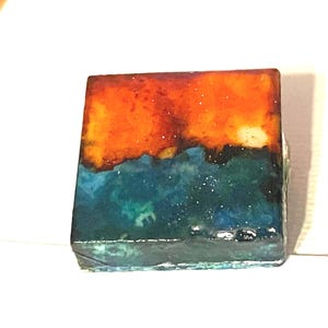 May include: Square art piece with a layered design. The top section is orange, transitioning to blue and green in the lower half. The piece has a glossy finish and appears to be made of resin or a similar material.