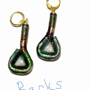 May include: A pair of drop earrings featuring gold-toned leverback hooks attached to a threaded metal post. The lower portion is a triangular shape with a metallic green and dark finish surrounding a central white triangular inlay. The word Backs is written in blue ink below the earrings.