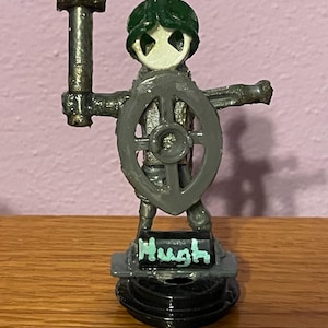 Könnte beinhalten: Eine kleine Metallfigur eines Kriegers mit grünem Helm und weißer Maske. Die Figur hält einen dunklen Hammer in der einen und einen grauen Schild in der anderen Hand. Ein schwarzer rechteckiger Sockel trägt den Namen Hugh in hellgrüner Farbe.