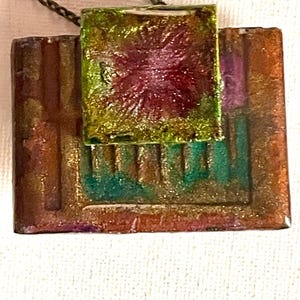 May include: A handcrafted pendant with a geometric design. The pendant features a square, textured, green and red center piece, and a rectangular base with vertical lines in shades of brown, orange, and green. It hangs from a dark chain.