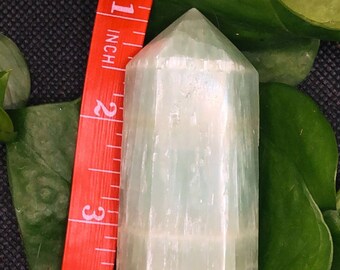 May include: A light green, white and orange crystal point, approximately 5 cm tall, is displayed next to a red measuring tape.