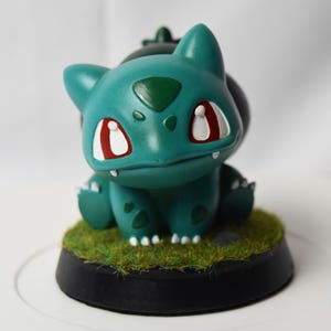 Puede incluir: Una figura de Bulbasaur color verde azulado con manchas verde oscuro, ojos blancos con pupilas rojas y pequeños colmillos blancos. La criatura se asienta sobre una base negra circular cubierta de césped artificial. Un logo de dragón azul es visible.