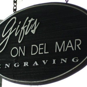 Może przedstawiać: Owalny, czarny drewniany szyld z białym napisem. Na szyldzie widnieje napis "Gifts ON DEL MAR ENGRAVING". Wisi na łańcuchu i prawdopodobnie służy do reklamowania sklepu z pamiątkami.