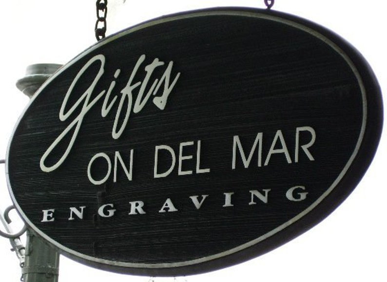Może przedstawiać: Owalny, czarny drewniany szyld z białym napisem. Na szyldzie widnieje napis "Gifts ON DEL MAR ENGRAVING". Wisi na łańcuchu i prawdopodobnie służy do reklamowania sklepu z pamiątkami.