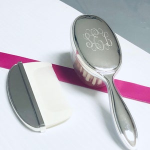 May include: A silver hairbrush and comb set with a monogrammed design. The brush has a white bristle head and a silver handle. The comb has a white plastic comb with a silver backing.