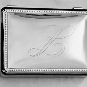 May include: A rectangular, silver-toned compact with a polished finish. The lid features an engraved cursive letter "L" and is framed by a beaded border. The edges are dark and reflective.