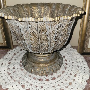May include: A decorative, antique-style pedestal bowl. The bowl features a scalloped rim and is adorned with a leaf pattern in gold and white. The bowl is set on a pedestal base, creating an elegant, vintage aesthetic.
