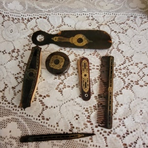 May include: A set of six vintage tortoiseshell and gold-accented grooming tools, including a comb, a brush, a small mirror, a razor, and two other tools. The tools are laid out on a white lace tablecloth.