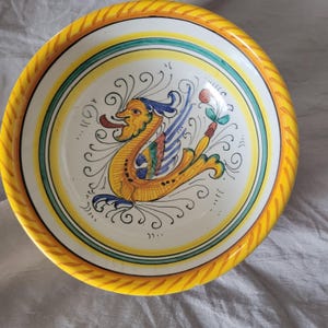 May include: A decorative ceramic bowl with a yellow rim and a detailed central design. The bowl features a yellow, blue, and green mythical creature design on a white background. The rim has a textured, rope-like pattern.